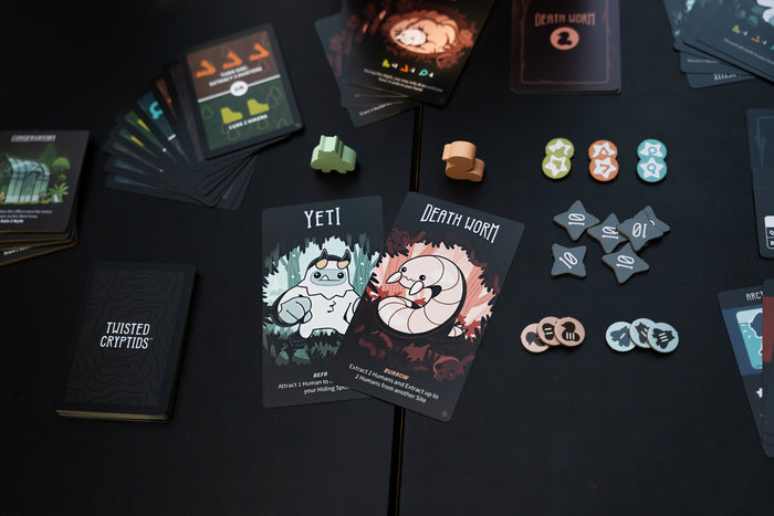 Cards, meeples and tokens from Twisted Cryptids Yeti or Not laid out on a black background. The center cards feature illustrations of the Yeti and the Death Worm.